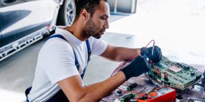 Arabian male auto mechanic examining a car engine removed from car for repair with voltmeter. Electronic Control Unit (ECU) of modern electric car.